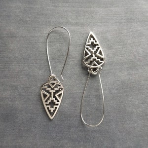 Puede incluir: Un par de pendientes plateados. Cada pendiente presenta un colgante en forma de lágrima con un diseño geométrico calado. Los pendientes tienen largos ganchos para la oreja curvados. El fondo es gris liso.