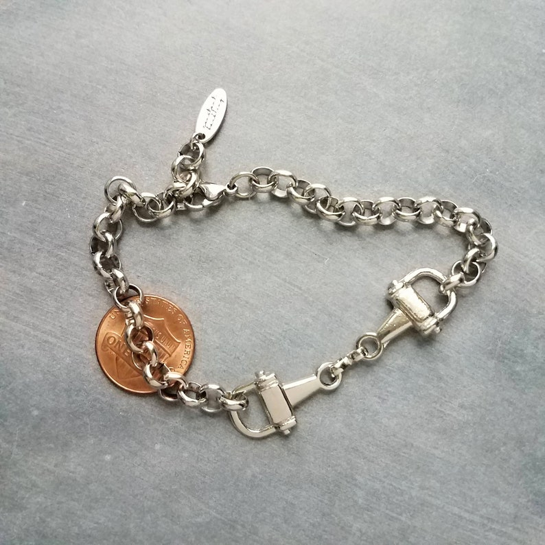May include: Silver-toned bracelet featuring a horse bit design. The bracelet has a chain-link style with a lobster clasp. A penny is next to the bracelet for size reference. The bracelet is on a gray surface.