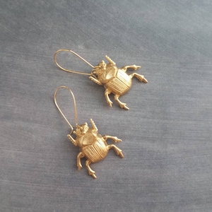Puede incluir: Pendientes dorados con forma de escarabajo. Cada pendiente presenta un diseño detallado y tridimensional de un escarabajo escarabajo con delicadas patas y antenas. Los pendientes tienen un cierre de gancho simple, y los escarabajos están orientados en direcciones opuestas.