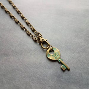 Può includere: Collana con ciondolo a forma di chiave in ottone antico. La chiave ha una patina turchese e il testo "922 H.CO". La collana ha una catena con chiusura a moschettone. Un accessorio vintage.