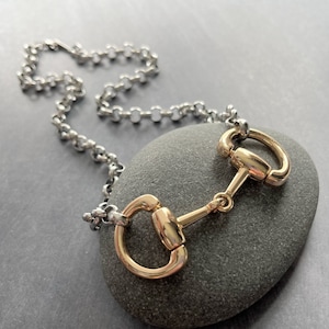 May include: A gold-toned equestrian bit pendant necklace with a silver-colored chain. The bit is centered on a gray stone, showcasing the jewelry's design. The necklace is a stylish accessory.