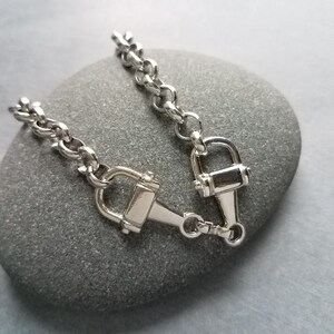 May include: A silver-colored equestrian-style bracelet featuring a horse bit design. The bracelet has a chain-link style with a clasp closure. The horse bit is approximately 1 inch wide.