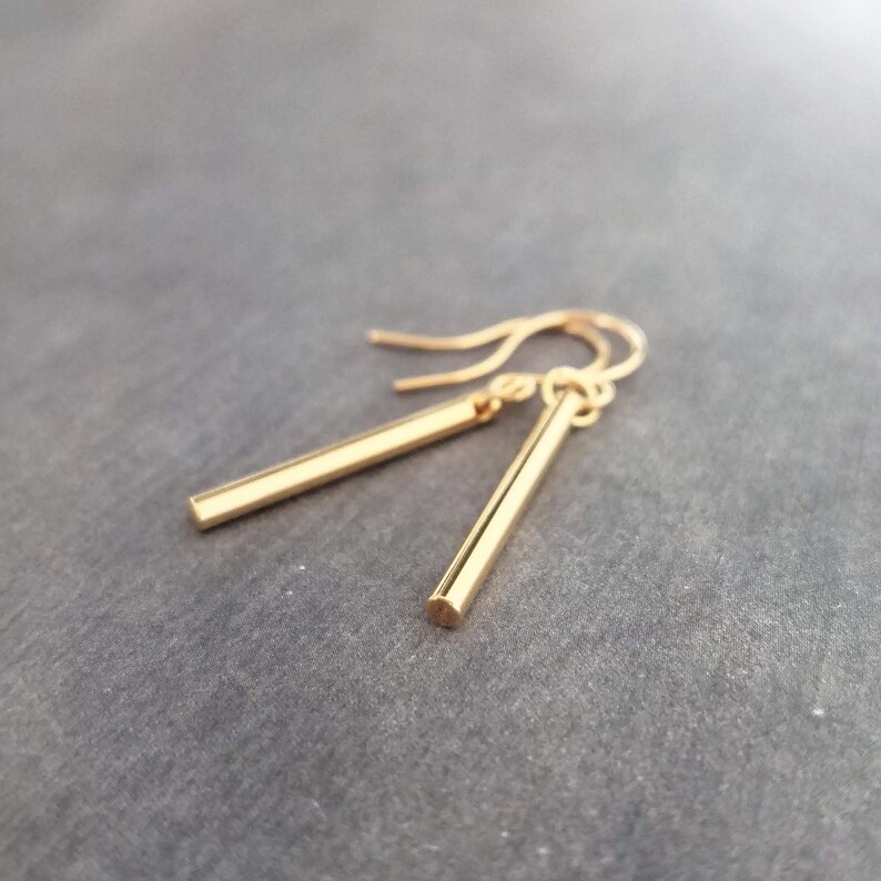 Peut inclure: Une paire de boucles d'oreilles pendantes en forme de barres dor&eacute;es. Chaque boucle d'oreille est compos&eacute;e d'une fine barre rectangulaire suspendue &agrave; un crochet simple. Les boucles d'oreilles sont pr&eacute;sent&eacute;es sur un fond gris.