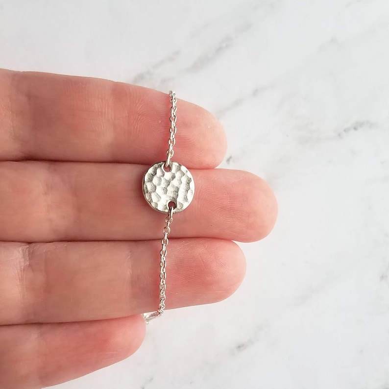 May include: A delicate silver bracelet with a hammered-texture, circular pendant. The pendant is connected to a fine chain. The bracelet is shown against a soft, neutral background, highlighting its simple elegance.