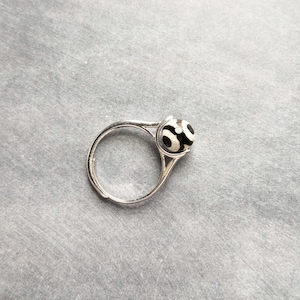 Puede incluir: Un anillo de plata con una cuenta estampada en blanco y negro. El anillo tiene un diseño simple y elegante con una banda redonda y un engaste que acuna la cuenta. La cuenta tiene un patrón distintivo en forma de ojo.