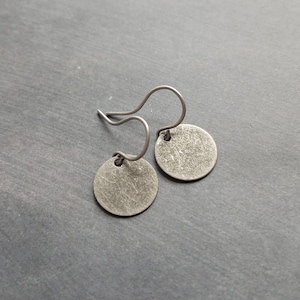 May include: A pair of silver-toned disc earrings with a textured surface. The earrings have a simple, minimalist design with a hook closure. The circular pendants are attached to the hooks with small loops. The background is a dark gray surface.