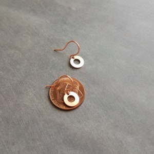 May include: A pair of small, copper-colored circle earrings with a hook closure. One earring is shown separately, and the other is placed on a penny for size comparison. The earrings have a minimalist design and a polished finish.