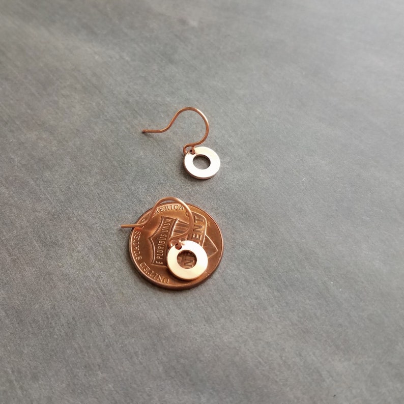 May include: A pair of small, copper-colored circle earrings with a hook closure. One earring is shown separately, and the other is placed on a penny for size comparison. The earrings have a minimalist design and a polished finish.