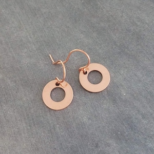 May include: A pair of copper-colored earrings. Each earring features a small, circular disc with a hole in the center, suspended from a curved ear wire. The earrings are displayed against a gray background.