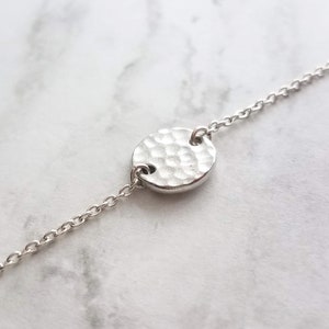 May include: A silver bracelet featuring a hammered-texture circular pendant. The pendant is linked to a delicate chain. The bracelet is shown on a white and grey marble surface. Jewellery.