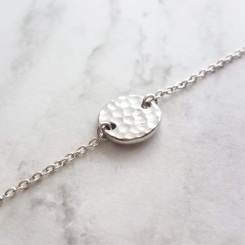 May include: A silver bracelet featuring a hammered-texture circular pendant. The pendant is linked to a delicate chain. The bracelet is shown on a white and grey marble surface. Jewellery.