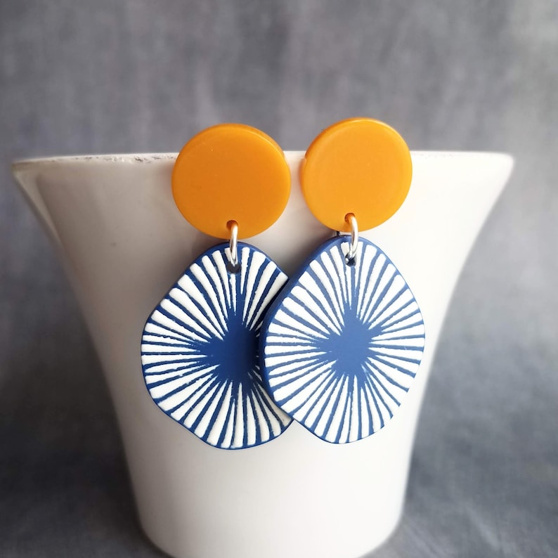 May include: A pair of earrings with a modern design. The earrings feature mustard yellow circular studs and navy blue, teardrop-shaped pendants with white radial patterns. The earrings are displayed against a white background.
