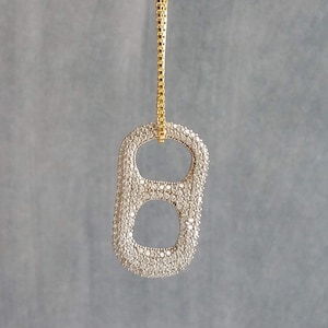 May include: A pendant necklace featuring a sparkling, silver-colored pop-top charm. The charm is encrusted with small, clear crystals and hangs from a delicate gold-colored chain. The pendant has two oval openings.
