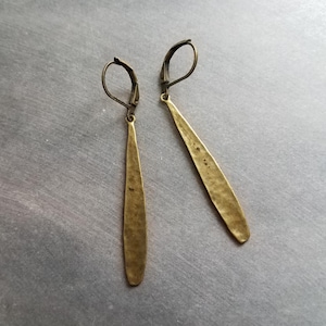 Op de afbeelding: Een paar lange, gehamerde messing oorbellen. Elke oorbel heeft een slanke, langwerpige druppelvorm, met een getextureerd oppervlak en een oorhaaksluiting. De oorbellen hebben een warme, antieke goudkleur.