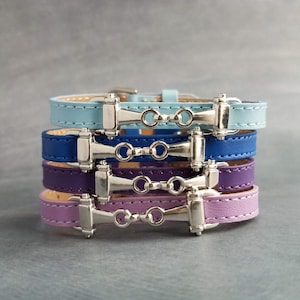 May include: Stack of four leather bracelets in shades of blue, purple, and lavender. Each bracelet features a silver-tone metal bit design. The bracelets are stacked on top of each other, showcasing the different colors and styles.