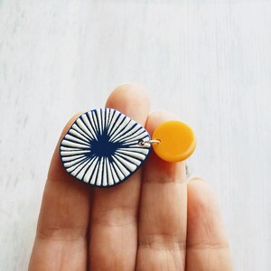 May include: A pair of asymmetrical earrings. One earring features a navy blue, square-shaped pendant with white radial lines. The other is a small, round, yellow pendant. Both are connected by a silver-colored metal loop.