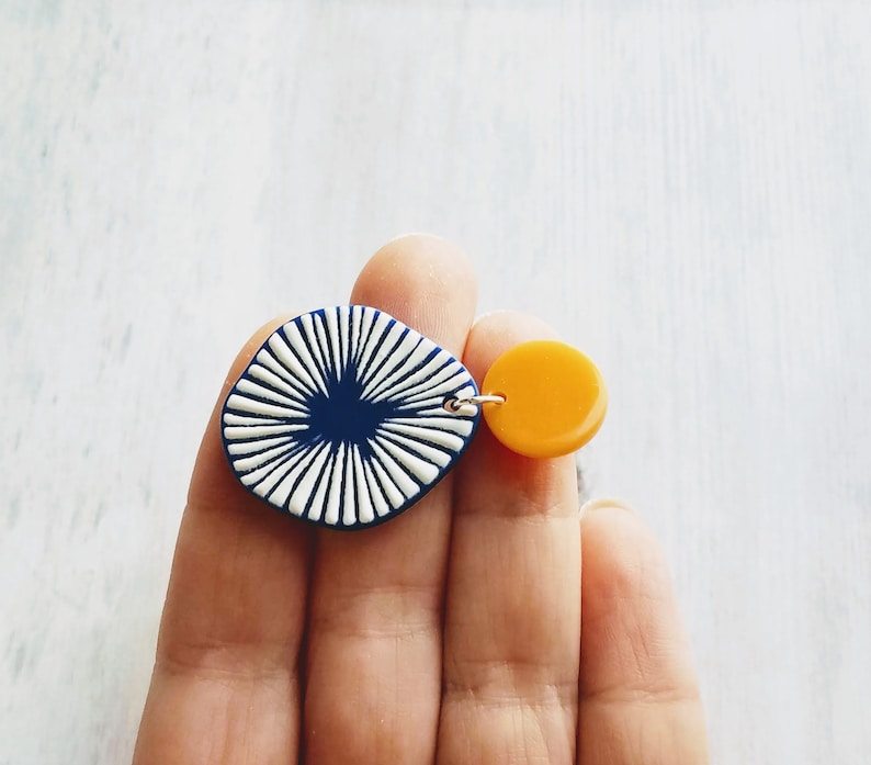 May include: A pair of asymmetrical earrings. One earring features a navy blue, square-shaped pendant with white radial lines. The other is a small, round, yellow pendant. Both are connected by a silver-colored metal loop.