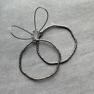 Puede incluir: Un par de pendientes de aro de metal gris oscuro martillado. Los aros tienen una forma irregular y orgánica y están suspendidos de sencillos ganchos para las orejas. Los pendientes se muestran sobre una superficie gris.