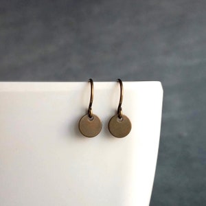 以下が含まれることがあります： 小さくて丸いブロンズ色のイヤリングのペア。各イヤリングは、湾曲したイヤーフックから吊り下げられた円形のペンダントが特徴です。イヤリングは、灰色の背景のある白い表面に表示されています。