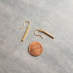 Peut inclure: Une paire de boucles d'oreilles en forme de barre dor&eacute;e avec fermoir crochet, pr&eacute;sent&eacute;es &agrave; c&ocirc;t&eacute; d'une pi&egrave;ce de monnaie pour comparaison de taille. Les boucles d'oreilles sont de conception simple et minimaliste, avec une finition polie.