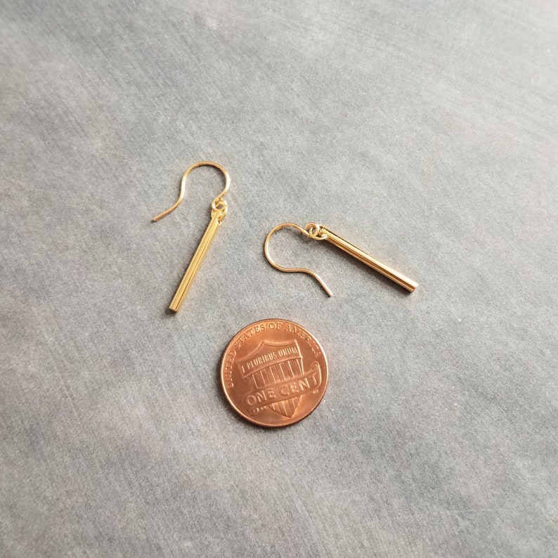 Peut inclure: Une paire de boucles d'oreilles en forme de barre dor&eacute;e avec fermoir crochet, pr&eacute;sent&eacute;es &agrave; c&ocirc;t&eacute; d'une pi&egrave;ce de monnaie pour comparaison de taille. Les boucles d'oreilles sont de conception simple et minimaliste, avec une finition polie.