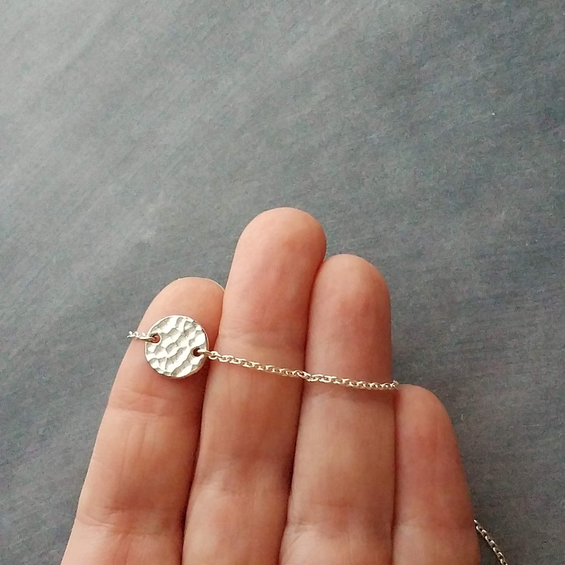 May include: A silver necklace with a hammered disc pendant. The pendant, with a textured surface, is round and attached to a fine chain. The necklace is shown against a grey background.