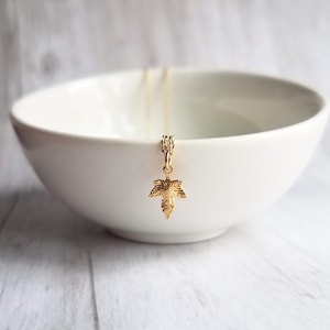May include: A delicate gold-colored necklace featuring a maple leaf pendant. The pendant is intricately detailed, showcasing the veins of the leaf. The necklace hangs in front of a white bowl, creating a simple yet elegant presentation.