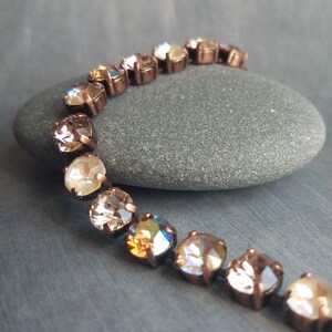 May include: A bracelet featuring a series of round, faceted crystals in shades of champagne and iridescent hues. The crystals are set in a copper-colored metal. The bracelet is displayed on a gray stone against a gray background.