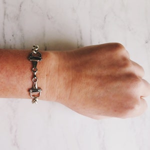 May include: Silver-toned equestrian-style bracelet. The bracelet features a series of connected bit-shaped links. The bracelet is displayed on a wrist against a white marble background. The bracelet is a stylish accessory.