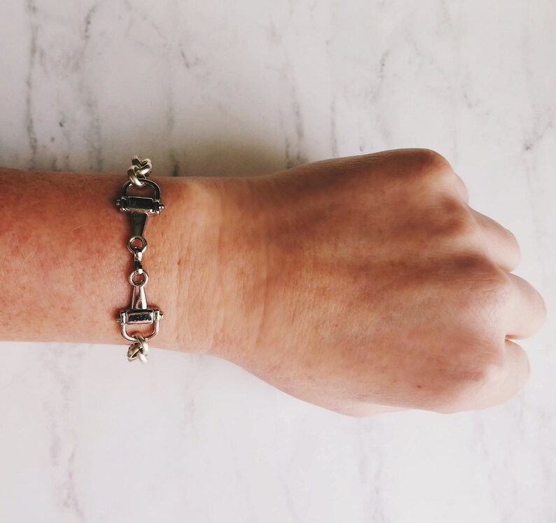 May include: Silver-toned equestrian-style bracelet. The bracelet features a series of connected bit-shaped links. The bracelet is displayed on a wrist against a white marble background. The bracelet is a stylish accessory.