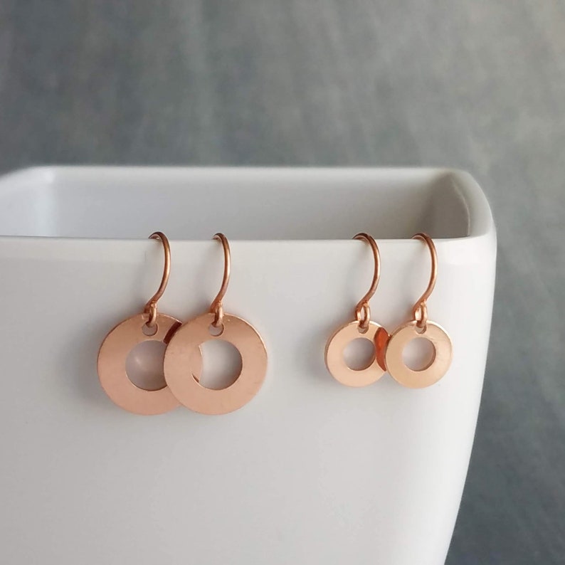May include: Rose gold circle earrings with a small circular cutout in the center. The earrings are displayed in a white container against a gray background. Two pairs of earrings are shown, one larger and one smaller.