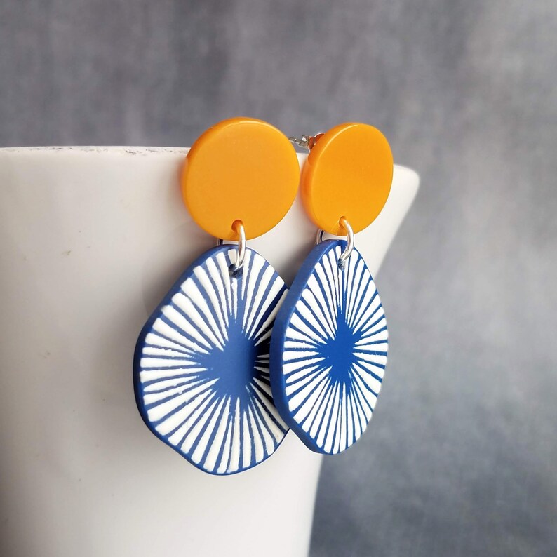May include: A pair of modern earrings featuring a bright orange circle stud and a blue teardrop pendant. The blue pendant has a white radial design. The earrings are hanging from a white ceramic container.