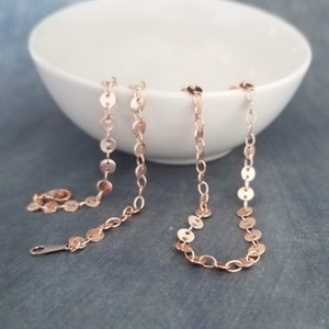 May include: Two delicate rose gold necklaces with a disc design are displayed against a white bowl and a grey background. One necklace is fully extended, showing the clasp, while the other is partially visible, showcasing the intricate chain.