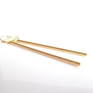 May include: A pair of gold-colored, linear drop earrings. Each earring features a long, slender bar suspended from a hook closure. The earrings are displayed against a white background, highlighting their simple, elegant design.