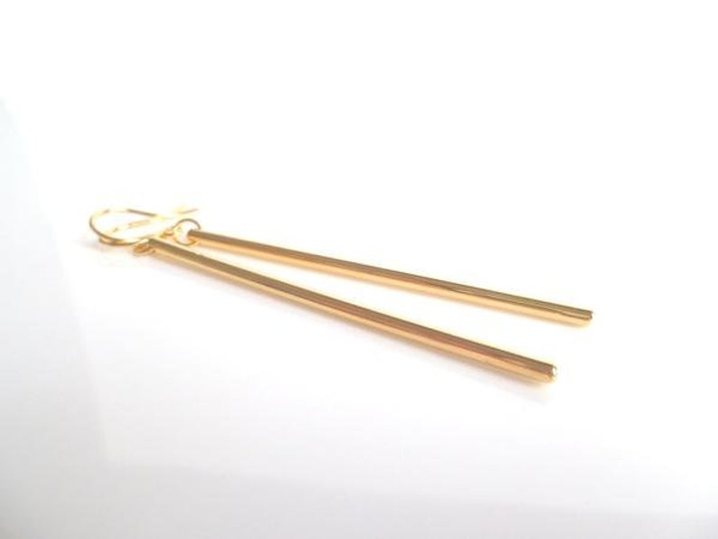 May include: A pair of gold-colored, linear drop earrings. Each earring features a long, slender bar suspended from a hook closure. The earrings are displayed against a white background, highlighting their simple, elegant design.