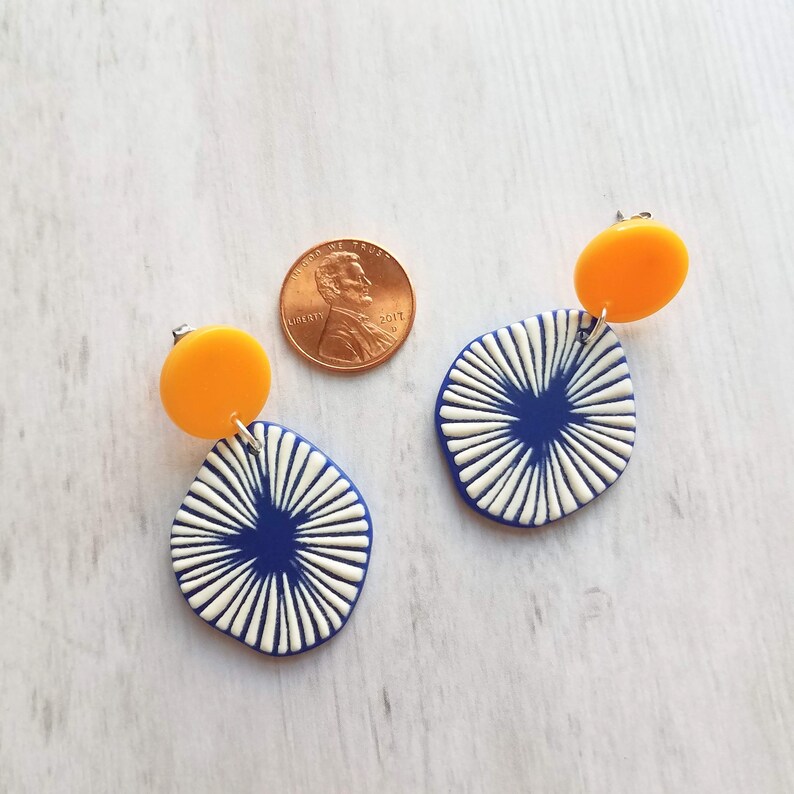 May include: A pair of modern earrings with a unique design. Each earring features a small, round, bright yellow stud, connected to a larger, abstract shape in navy blue with white radial lines. A penny is included for size reference.