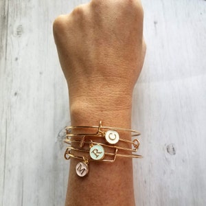 Puede incluir: Pulseras de brazalete doradas con dijes de iniciales. Las pulseras están apiladas en una muñeca, con dijes que muestran las letras "M", "R" y "C" en esmalte rosa, celeste y blanco. El fondo es un veteado de madera clara.
