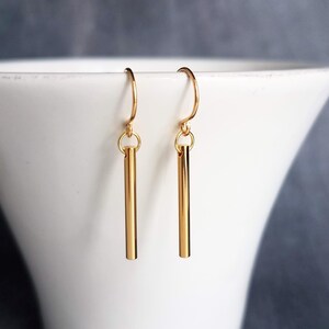 Peut inclure: Une paire de boucles d'oreilles dor&eacute;es en forme de barres. Chaque boucle d'oreille est compos&eacute;e d'une barre droite suspendue &agrave; un crochet incurv&eacute;. Les boucles d'oreilles sont pr&eacute;sent&eacute;es sur fond blanc.