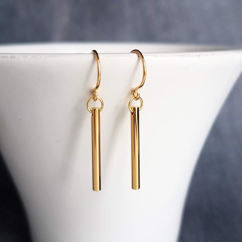Peut inclure: Une paire de boucles d'oreilles dor&eacute;es en forme de barres. Chaque boucle d'oreille est compos&eacute;e d'une barre droite suspendue &agrave; un crochet incurv&eacute;. Les boucles d'oreilles sont pr&eacute;sent&eacute;es sur fond blanc.