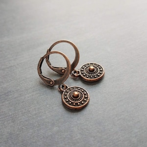 May include: A pair of copper-colored leverback earrings. Each earring features a circular pendant with a central raised design surrounded by a dotted pattern. The earrings have a vintage or antique aesthetic.