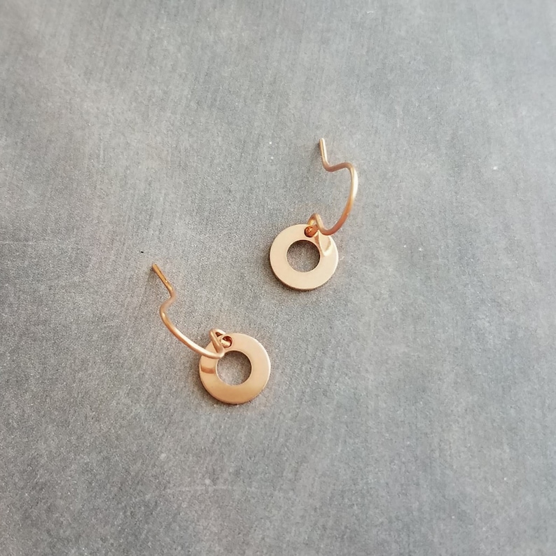 May include: Two rose gold hoop earrings with a circular pendant. The earrings have a simple, minimalist design. The earrings are displayed on a gray surface.