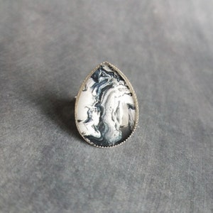 May include: A teardrop-shaped ring with a silver-colored band. The ring's face features a swirling pattern of white, black, and gray, resembling marble. The ring is set against a gray background.