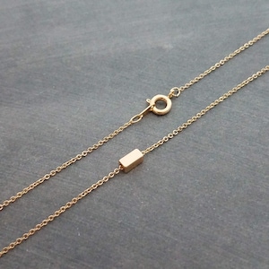 以下が含まれることがあります： 小さな長方形のペンダントが付いた繊細なゴールドネックレス。チェーンは細く、留め金が見えます。ネックレスは濃い灰色の背景に表示されています。