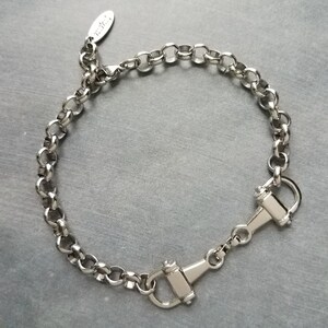 May include: A silver-colored bracelet with a chain-link design. The bracelet features a clasp closure with a hook and a small tag with text. The bracelet is displayed on a gray surface.