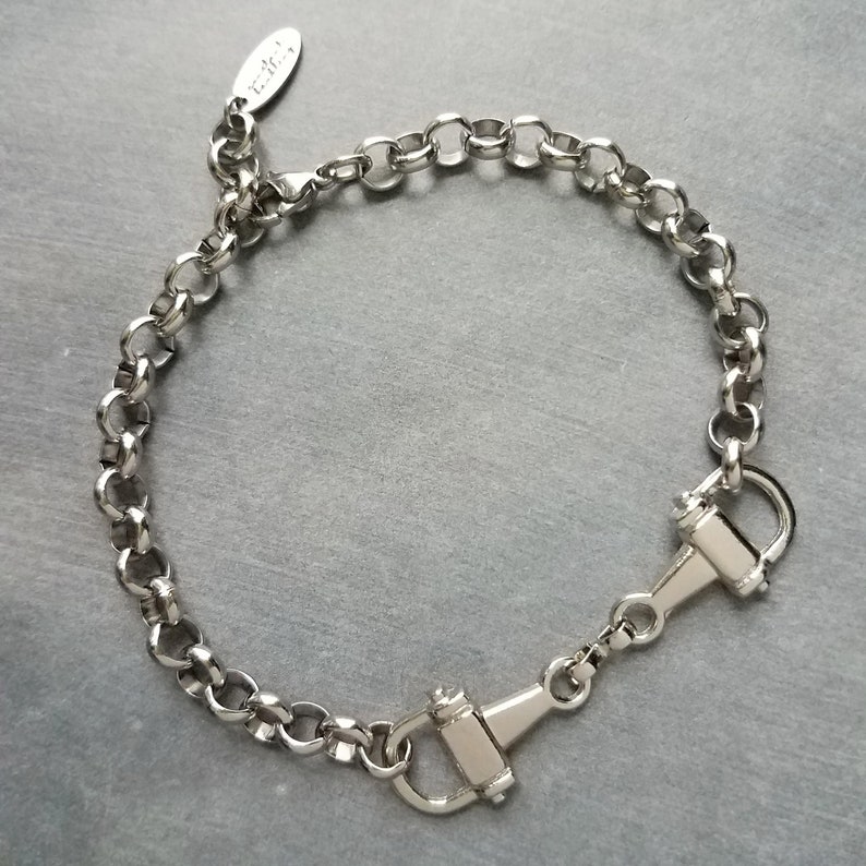 May include: A silver-colored bracelet with a chain-link design. The bracelet features a clasp closure with a hook and a small tag with text. The bracelet is displayed on a gray surface.