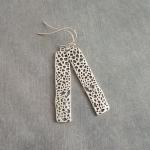 Könnte beinhalten: Ein Paar silberfarbene Ohrhänger. Jeder Ohrring hat eine lange, rechteckige Form mit unregelmäßigen Kanten und einem Muster aus kleinen, unregelmäßigen Löchern. Die Ohrringe hängen an einem einfachen Haken.