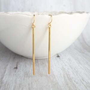 May include: A pair of gold-colored bar earrings. The earrings are long, slender, and hang from a simple hook. The earrings are displayed against a white background.
