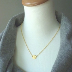 May include: A delicate gold necklace with a small, textured circular pendant. The pendant hangs from a fine chain, displayed on a mannequin. The background is a soft grey, highlighting the jewelry's simple elegance.