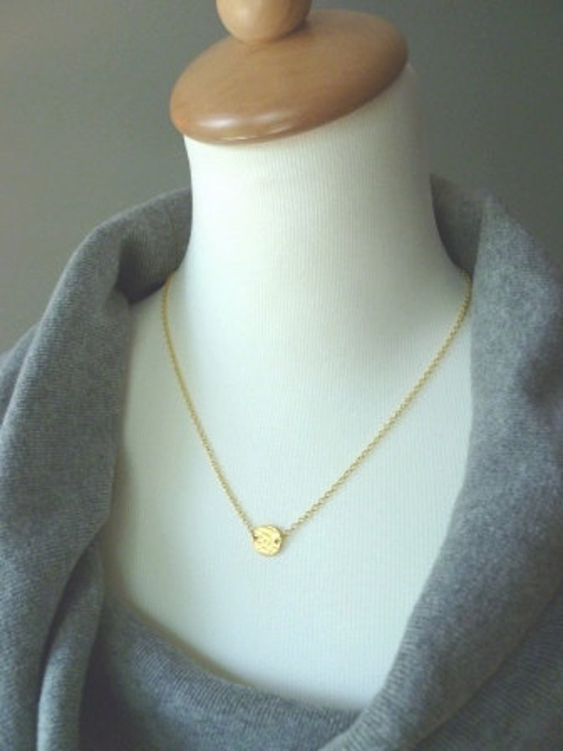 May include: A delicate gold necklace with a small, textured circular pendant. The pendant hangs from a fine chain, displayed on a mannequin. The background is a soft grey, highlighting the jewelry's simple elegance.