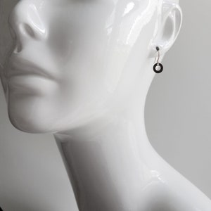 May include: A single black circle earring hangs from a mannequin's ear. The earring is a simple, minimalist design, with a small, dark circle suspended from a thin hook. The mannequin is white and glossy, with a smooth surface.